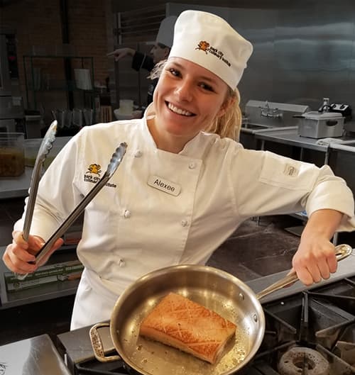 A Park City Culinary Institute student pan-frying salmon and holding tongs while smiling.