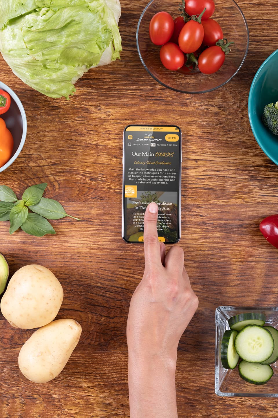 The Park City Culinary Institute's homepage displayed on a mobile phone on a kitchen counter, surrounded by cooking ingredients.